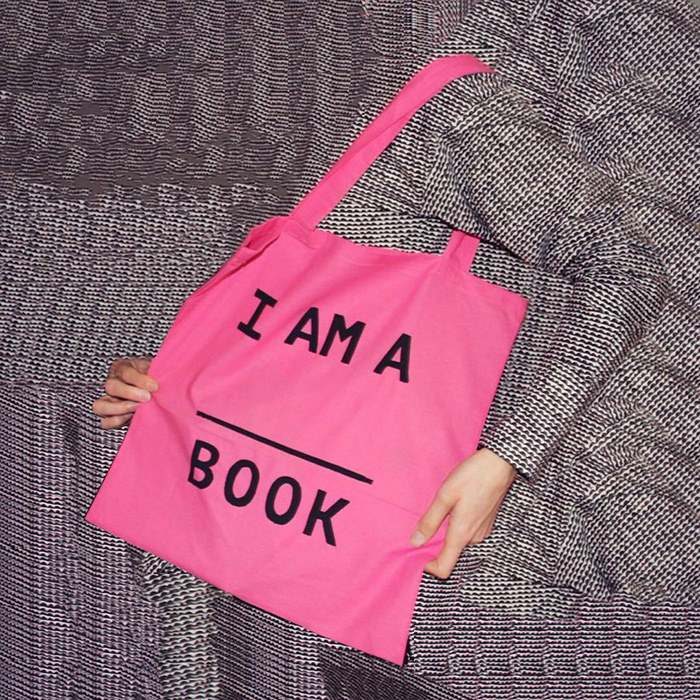 A black and white shirt expands to a square, an arm with a bright pink tote is at the center, the tote says 'I am a ______ book'