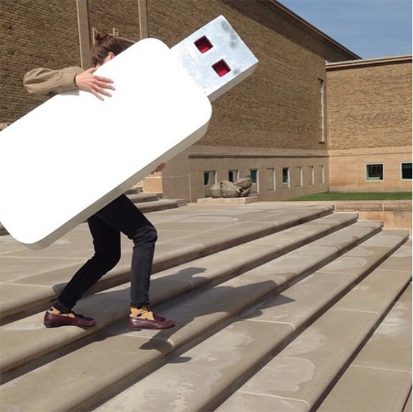 a person carries a larger than life (human-sized) thumbdrive down beige stairs